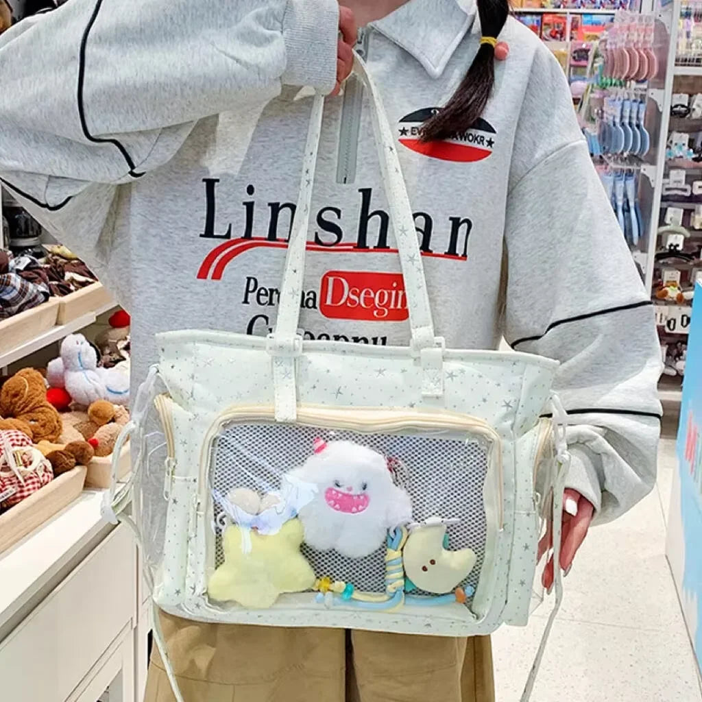 A person holds a transparent white shoulder tote bag with a pink and yellow cloud charm in a retail setting.