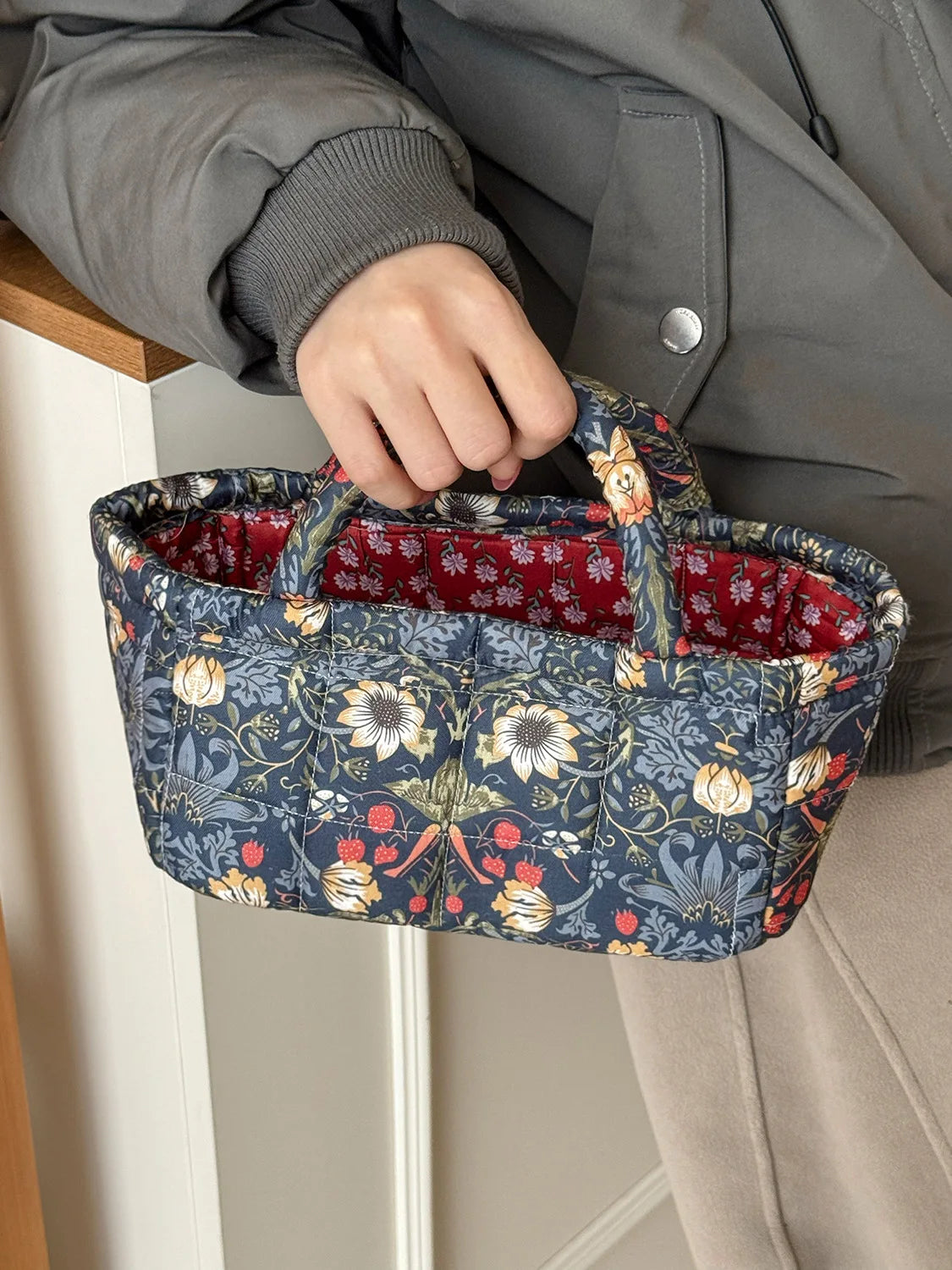 A hand holds a quilted fabric storage basket with blue floral embroidery against a gray jacket.