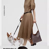 A woman wearing a beige shirt and skirt holds a brown geometric embroidery hobo shoulder bag while a dog stands beside her.