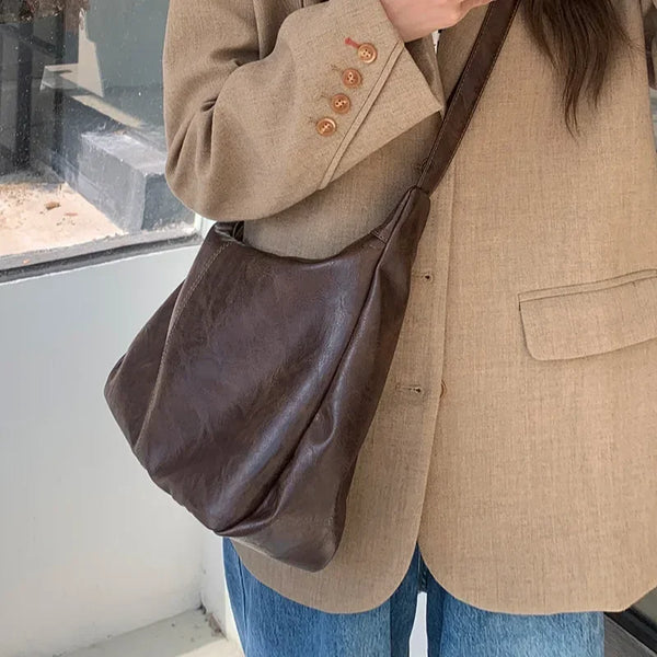 A person is holding a brown vintage PU leather tote bag with a long strap against a light colored jacket and blue jeans.