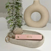 A pink leather wristlet keychain with the name Olivia rests on a light beige woven tray next to a smooth ceramic ring.