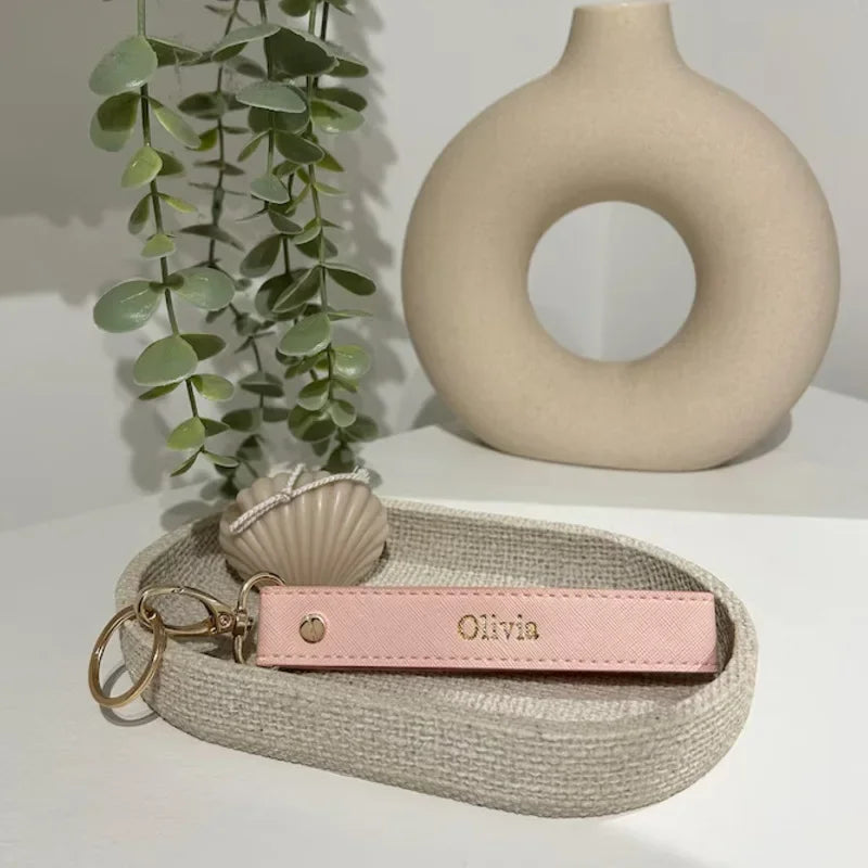 A pink leather wristlet keychain with the name Olivia rests on a light beige woven tray next to a smooth ceramic ring.