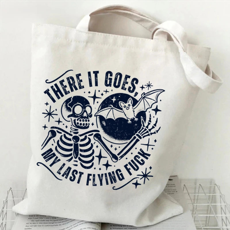 A white canvas tote bag featuring a black ink design of a skull and bat flying over a pumpkin with text.