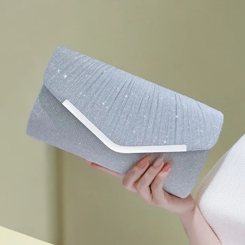 A hand holds a light gray glitter pleated crossbody clutch bag with a white trim against a pale background.
