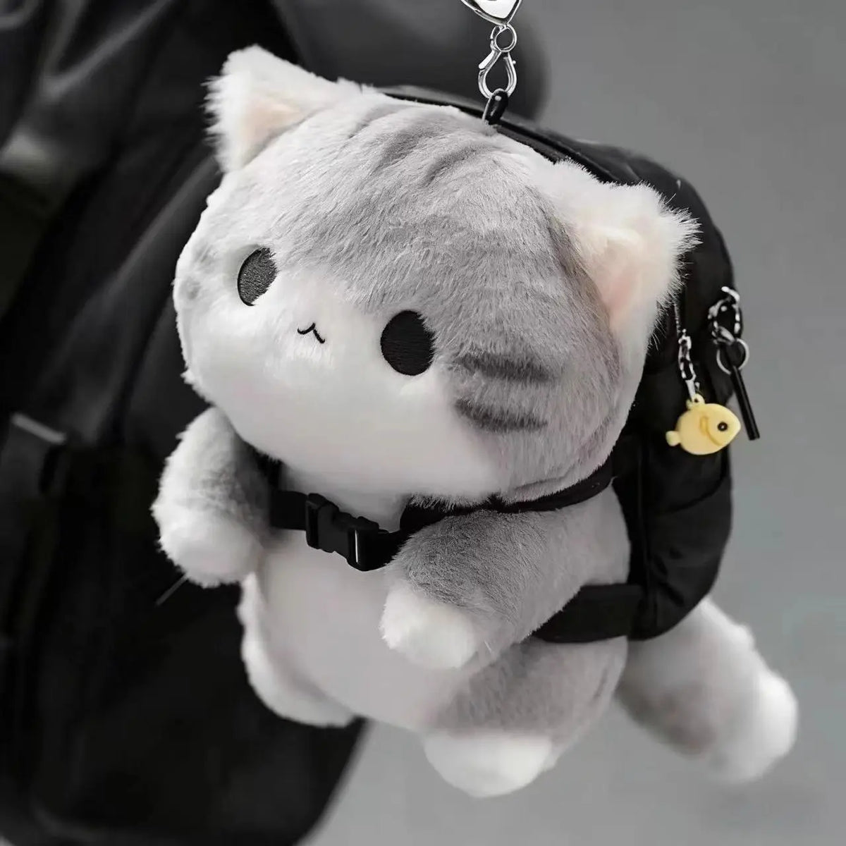 A grey soft cat plush pendant attached to a black backpack with a small charm.