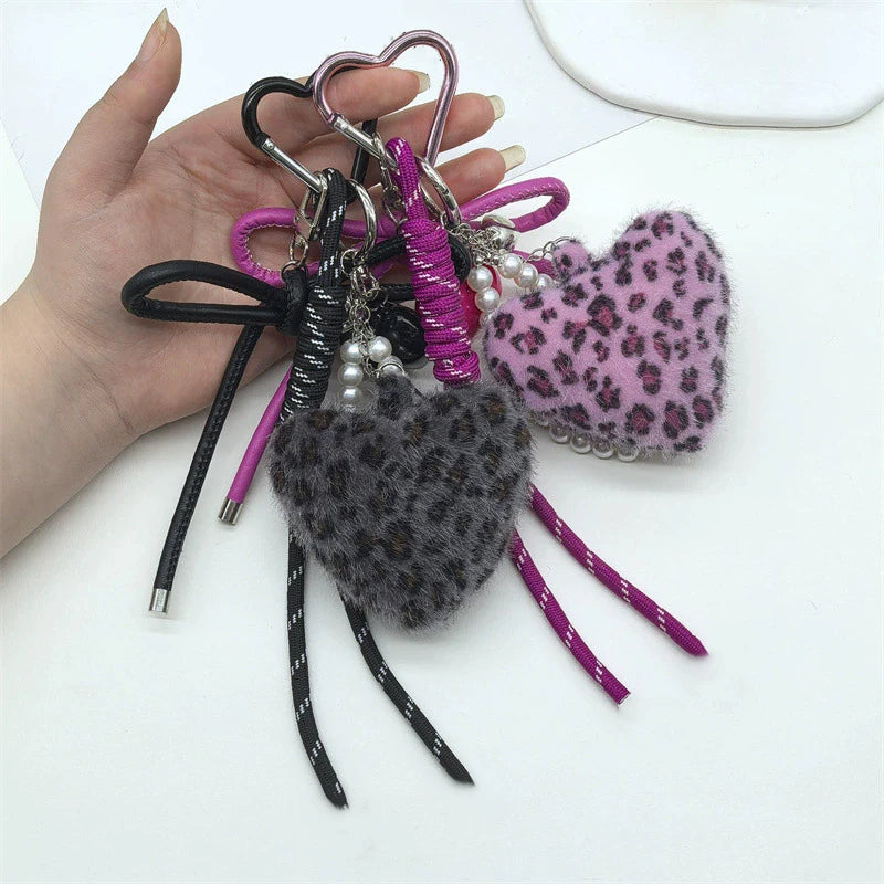 A hand holds a collection of heart-shaped charm keychains featuring black, purple, and pink leopard print faux fur.