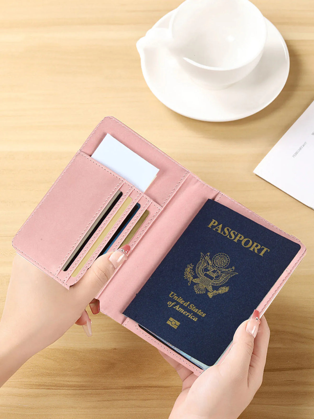 A hand holds a pink slim passport holder containing a blue passport and an ID card on a wooden table.