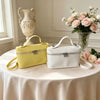 A yellow and white minimalist pebbled leather box crossbody bag displayed on a table with floral arrangements.