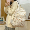 Una persona sostiene un suéter de punto de color crema y lleva una mochila con estampado floral y múltiples bolsillos.