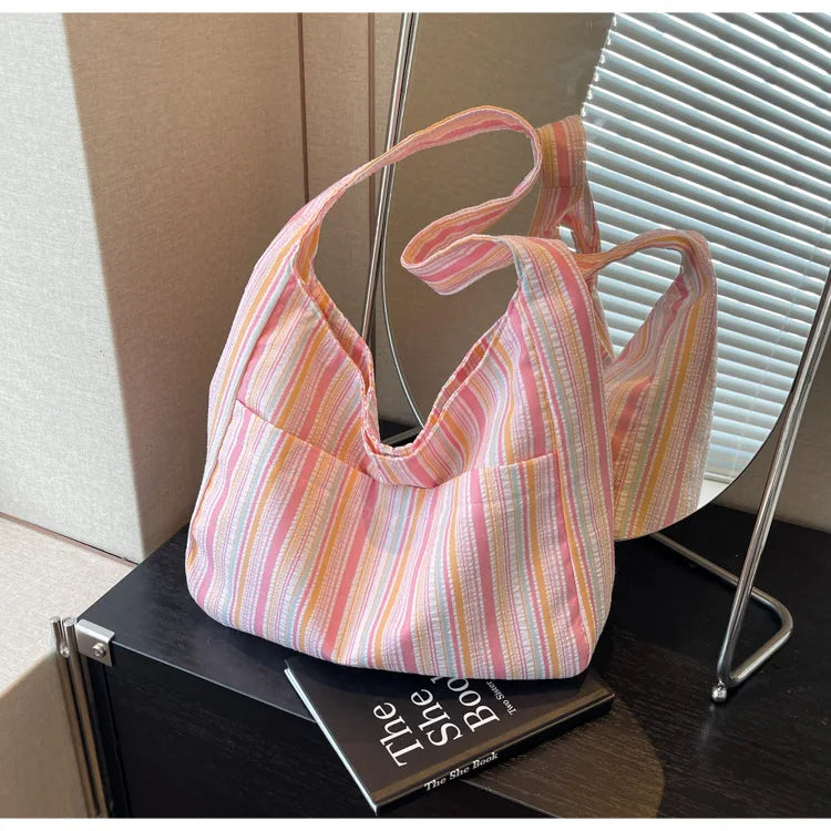 A pink and white striped canvas shoulder bag is displayed on a dark surface next to a book.