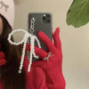 A hand wearing a red sweater holds a smartphone displaying a pearl bowknot keychain with a heart charm.