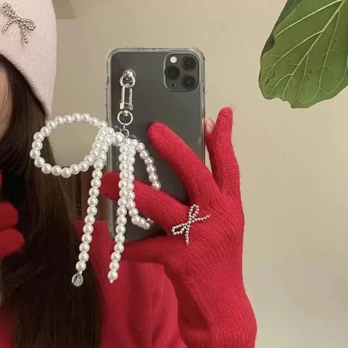 A hand wearing a red sweater holds a smartphone displaying a pearl bowknot keychain with a heart charm.