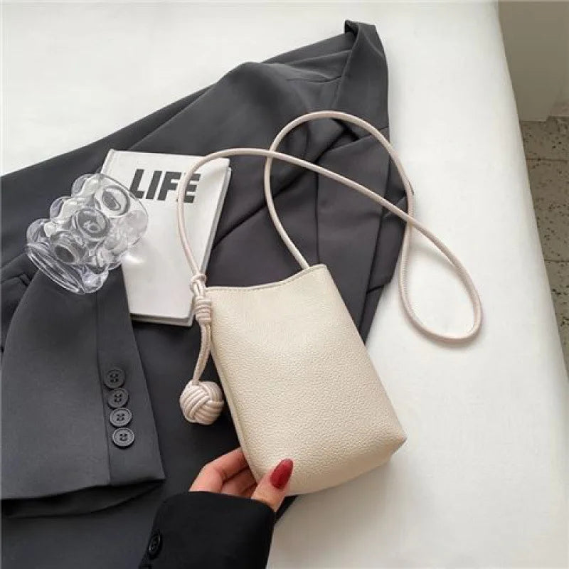 Bolso cruz de cuero beige con correa rosa claro junto a un blazer gris y una tarjeta blanca.