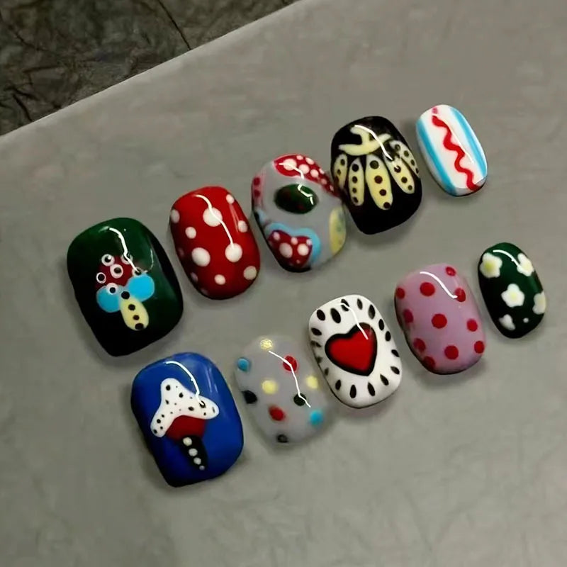 A collection of 10 colorful, cartoon-themed press-on nails shaped like beans and oval tips arranged on a light surface.