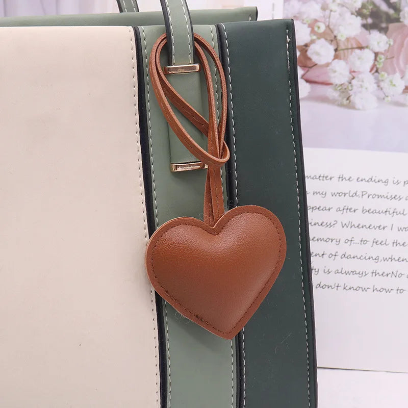 A brown leather heart keychain charm attached to a green and beige vertical leather bag strap.