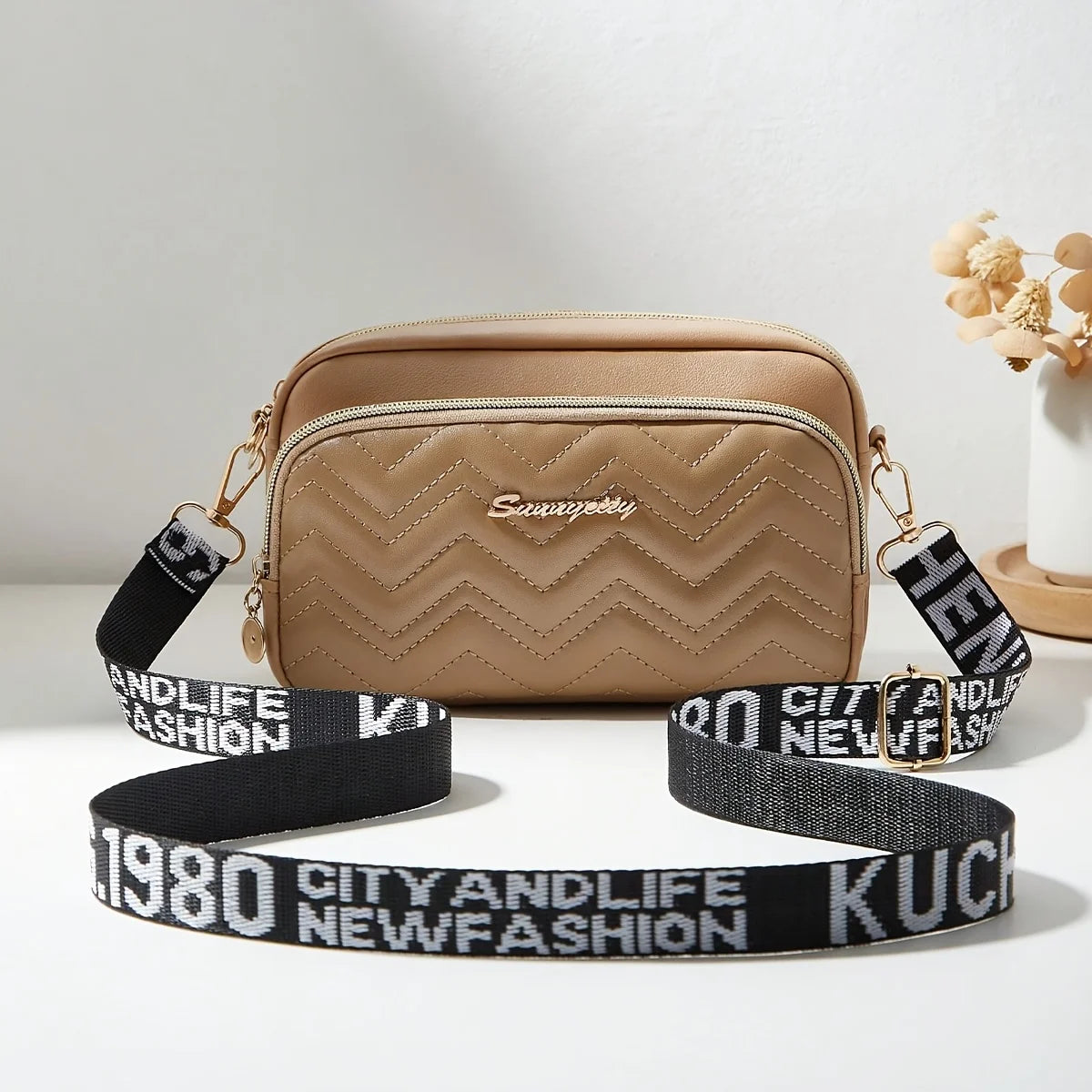 Beige quilted crossbody camera bag with a black letter strap resting on a white surface.
