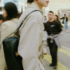 A person wearing a beige trench coat and carrying a black leather square shoulder crossbody bag on a city street.
