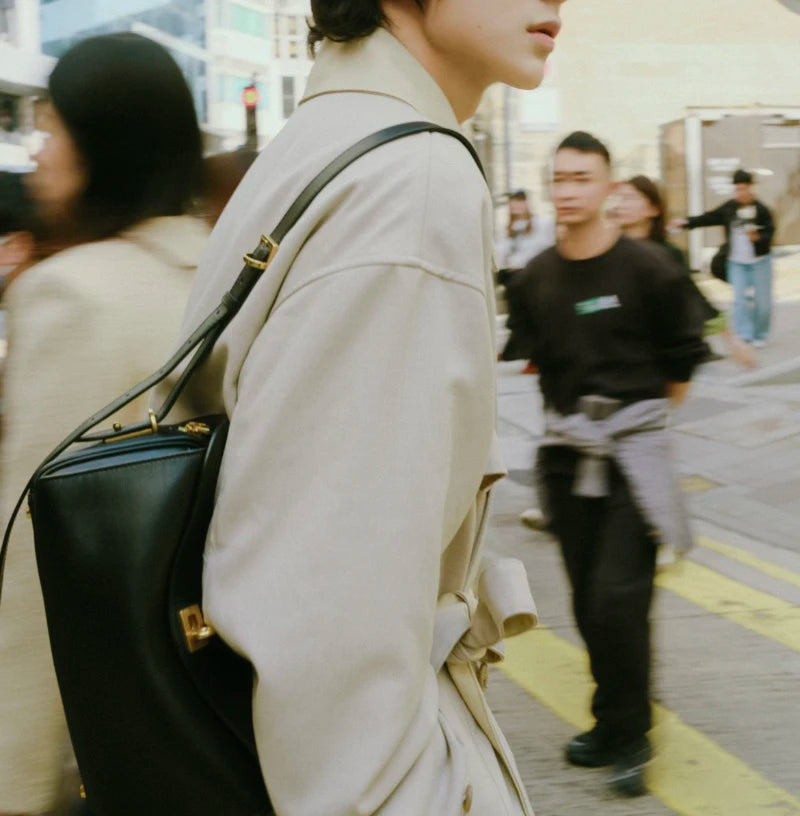 A person wearing a beige trench coat and carrying a black leather square shoulder crossbody bag on a city street.