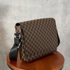 A brown patterned flap crossbody bag resting on a wooden stool against a gray background.