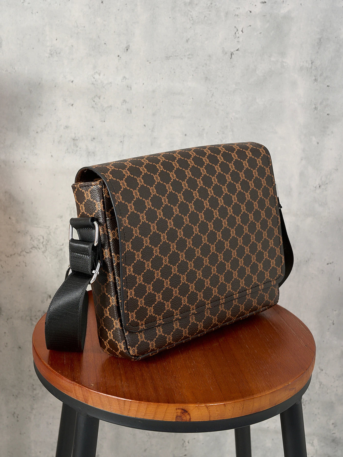 A brown patterned flap crossbody bag resting on a wooden stool against a gray background.