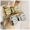 A close-up of a gold and silver bowknot evening bag with detachable chain straps resting on a light beige fabric.