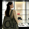 A woman wearing a gold sequined dress holds a large olive green knitted tote bag with a dark strap in front of a glass displa