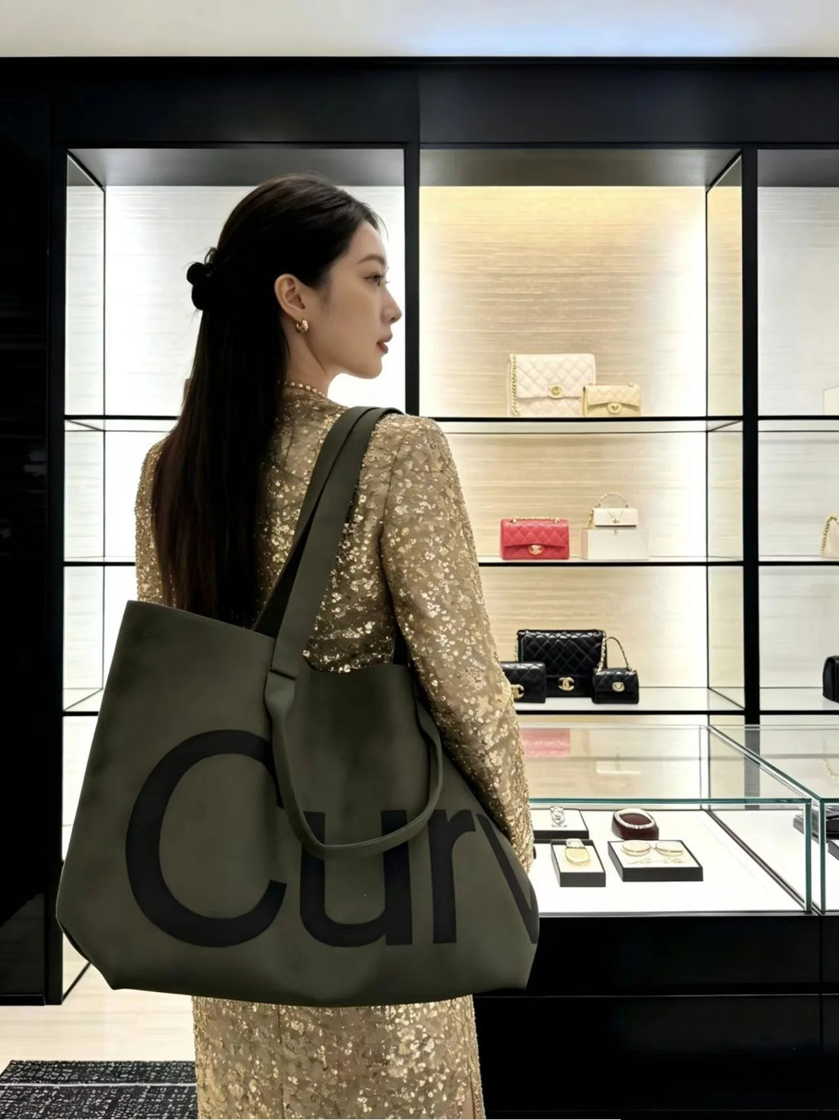 A woman wearing a gold sequined dress holds a large olive green knitted tote bag with a dark strap in front of a glass displa