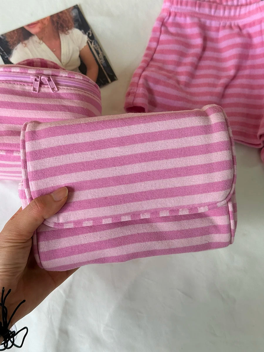 A hand holds a pink striped makeup bag with a flip-top mirror, next to pink striped shorts.