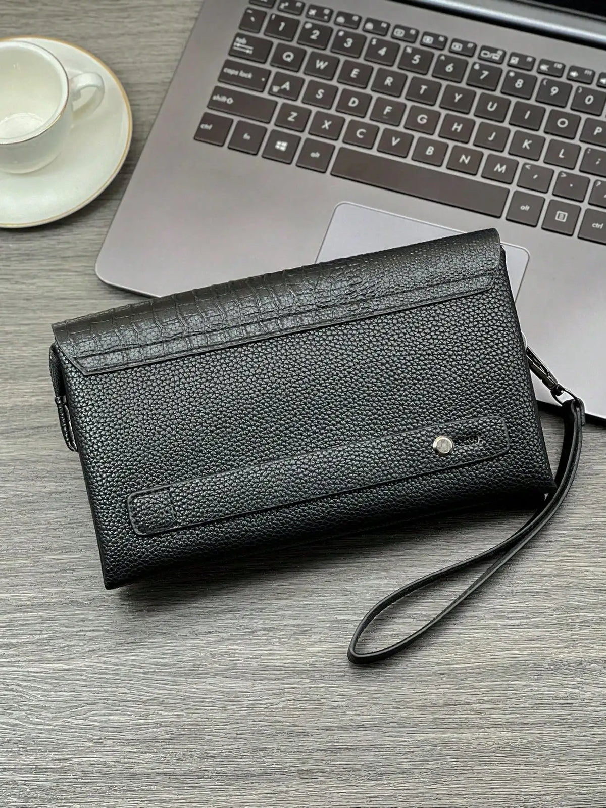 A black crocodile pattern clutch bag resting on a wooden desk next to a silver laptop and a white cup.