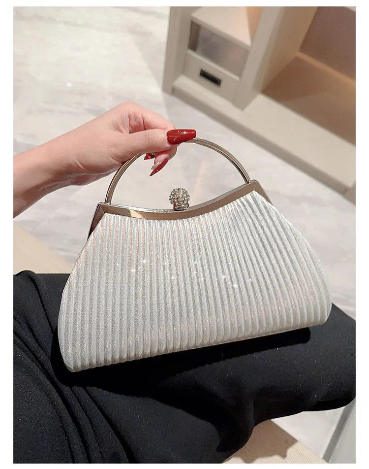 A hand holds a light silver pleated glitter evening handbag with a top handle and chain strap against a white background.
