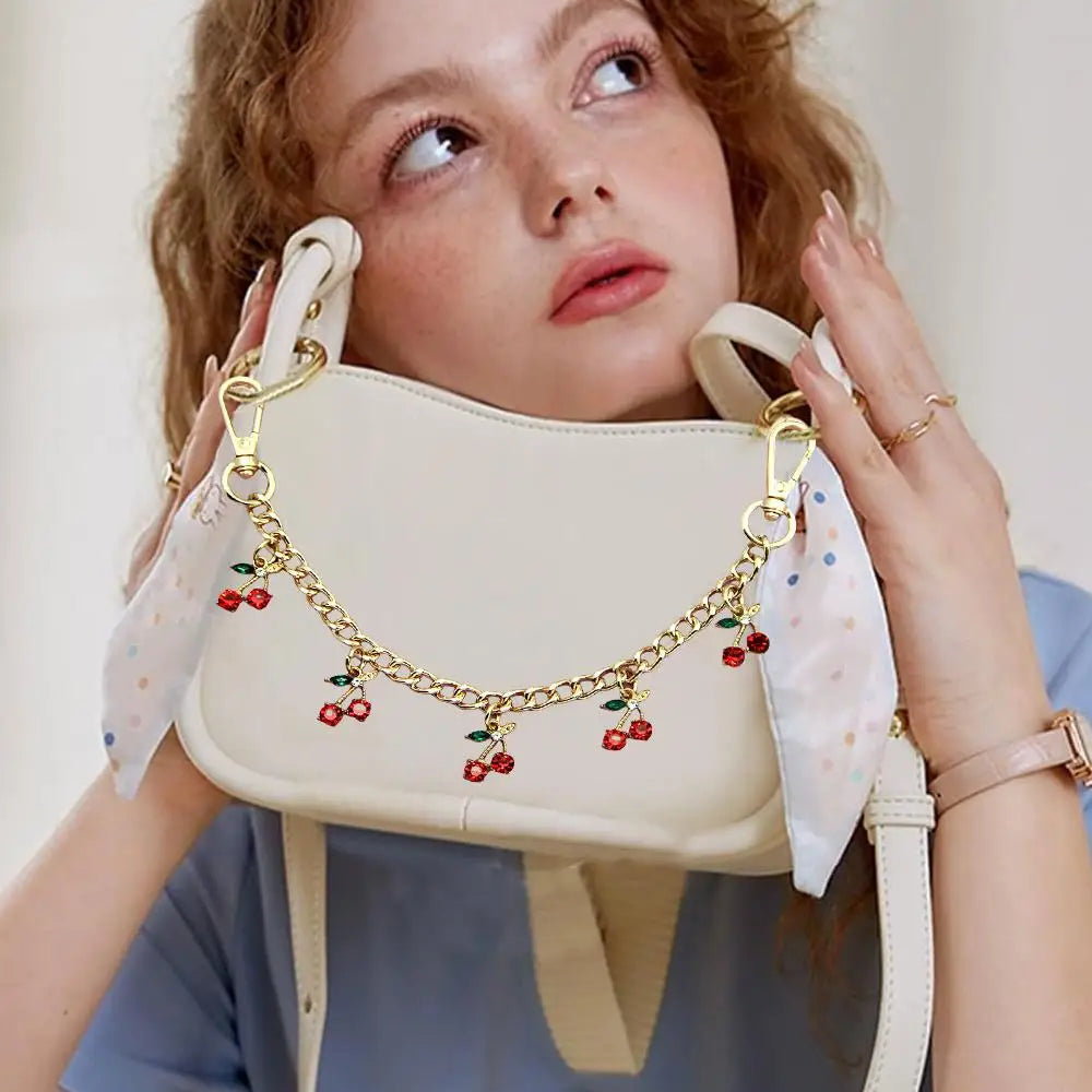 A woman holds a light beige bag decorated with gold chain pendants featuring small red berries and leaves.