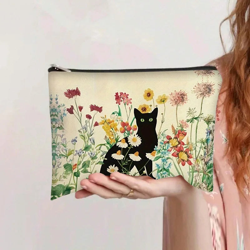 Una mano sostiene una bolsa de cosméticos beige con estampado floral de gatos negros y diversas flores silvestres coloridas.