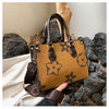 A tan leather shoulder tote bag with dark brown floral patterns rests on a pile of gray rocks.