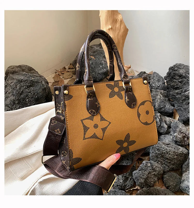 A tan leather shoulder tote bag with dark brown floral patterns rests on a pile of gray rocks.