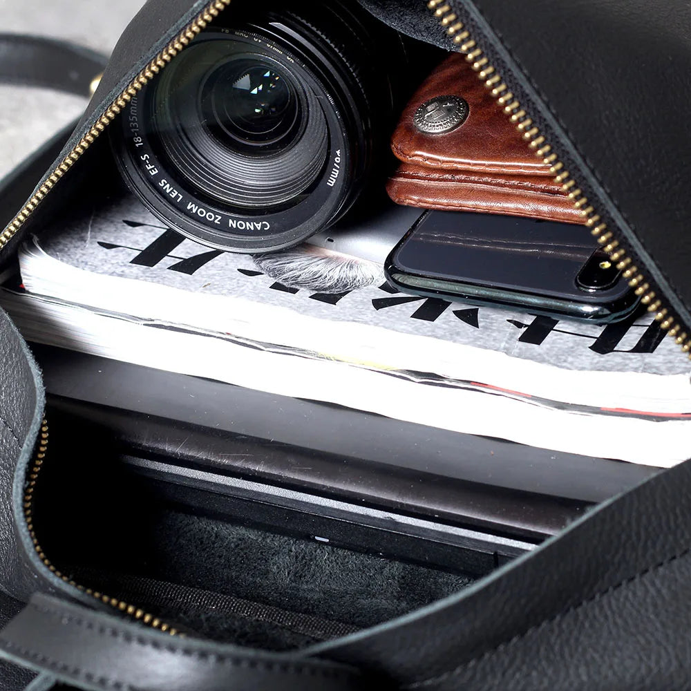 A close-up view inside a black leather backpack showing a camera lens, a smartphone, and some paper.