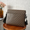 A brown patterned flap crossbody bag resting on a wooden stool with a vase of pink flowers in the background.