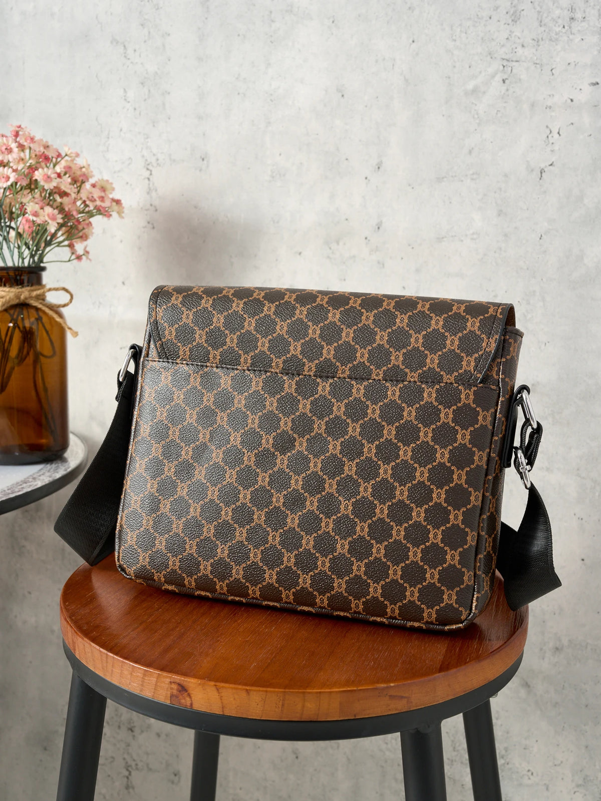 A brown patterned flap crossbody bag resting on a wooden stool with a vase of pink flowers in the background.