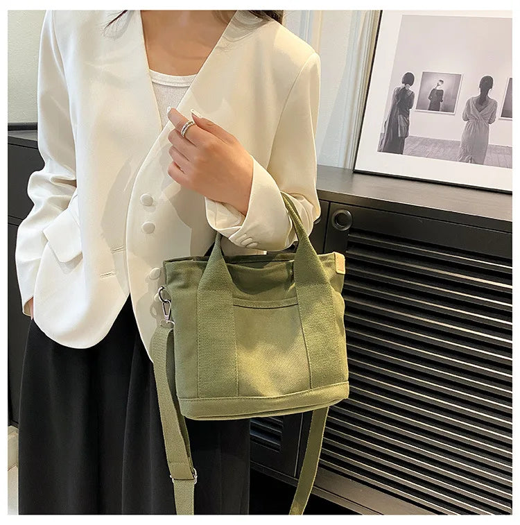 A woman models a light green canvas crossbody bag with an adjustable strap and multiple pockets against a dark background.