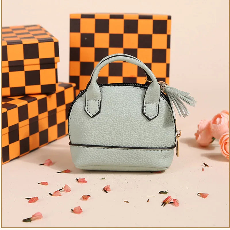A light mint green portable earbud case with a black trim and a tassel charm is displayed next to a checkered orange gift box