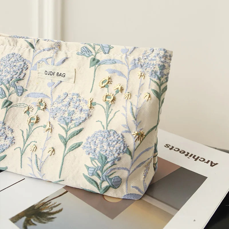 A light beige floral canvas makeup bag featuring blue and green botanical embroidery.