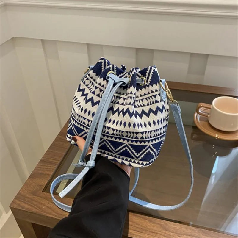 A navy and cream patterned drawstring bucket bag with light blue straps resting on a glass surface.