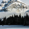 Majestätischer schneebedeckter Berggipfel umgeben von Nebel und einem dunklen Nadelwald auf einer ruhigen Wasserfläche.