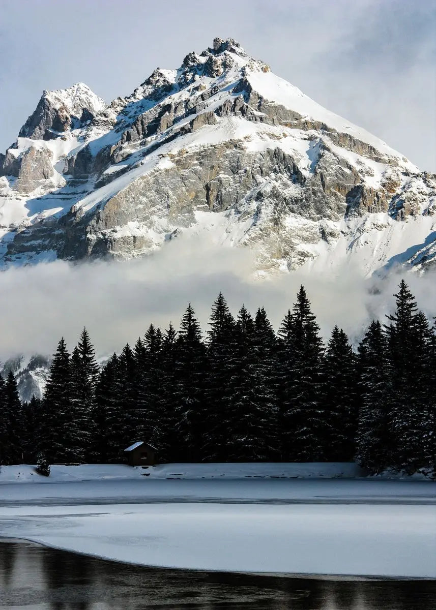 Majestätischer schneebedeckter Berggipfel umgeben von Nebel und einem dunklen Nadelwald auf einer ruhigen Wasserfläche.