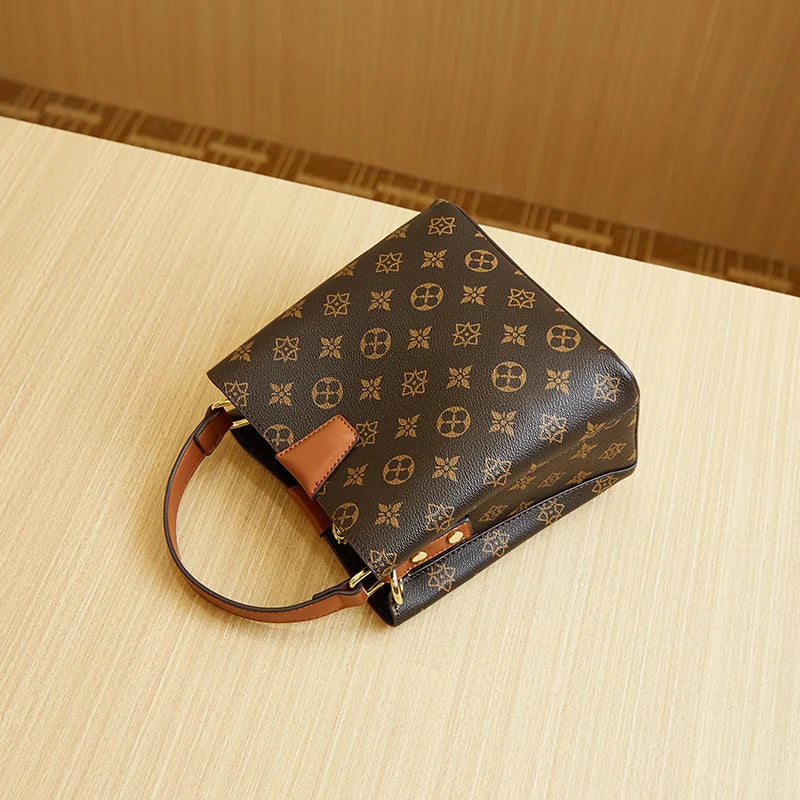A dark brown leather bucket bag with gold accents and a tan adjustable crossbody strap resting on a light wood surface.