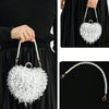 A close-up of a white, heart-shaped handbag covered in silver pearls, held by a woman against a black background.