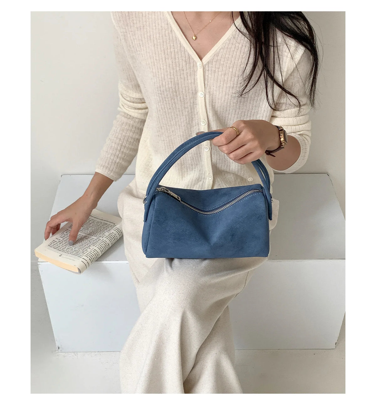 A woman wearing a cream knit sweater holds a navy blue faux suede crossbody bag.