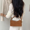 A woman is shown holding a tan faux suede crossbody bag against a light background.