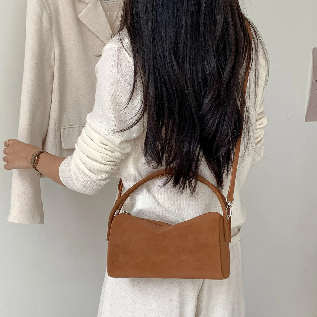 A woman is shown holding a tan faux suede crossbody bag against a light background.