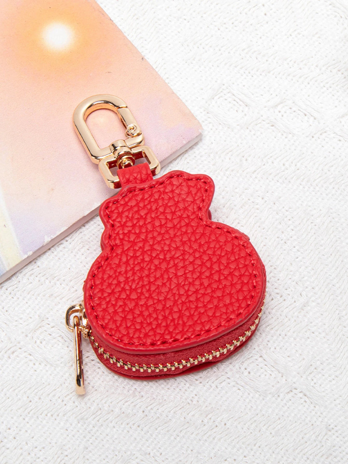 Cartera de monedas con forma de panda roja y texturizada, con cierre y cadena dorada sobre fondo rosa claro.