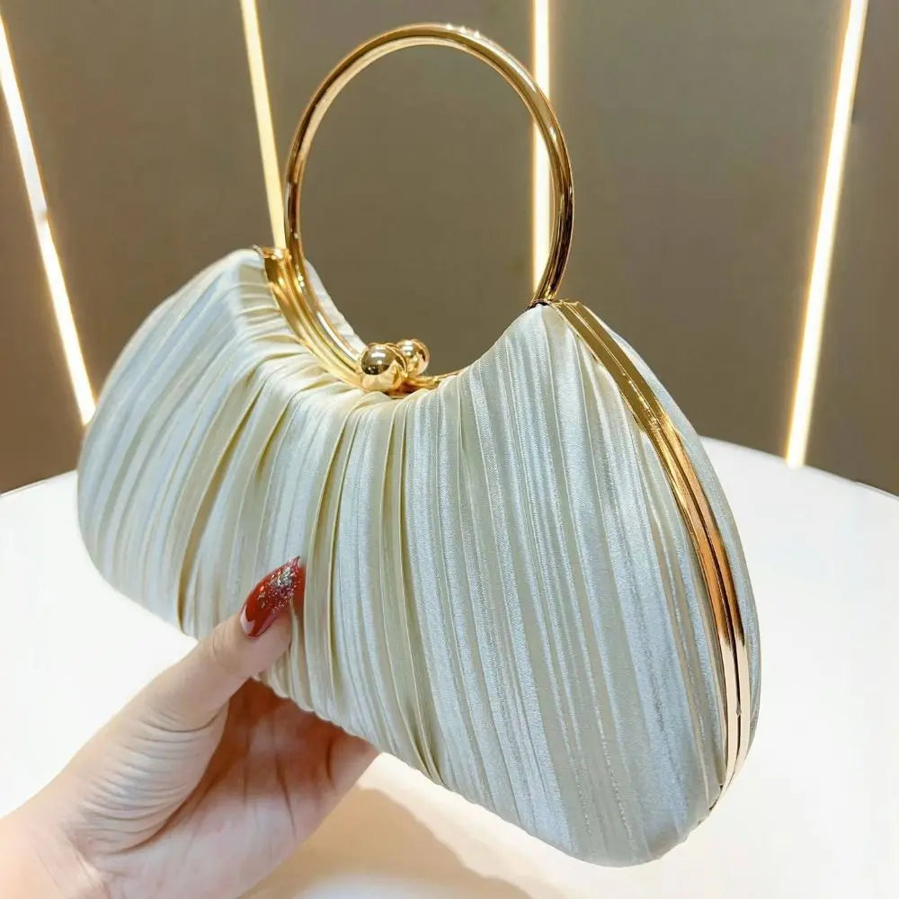 A hand holds an elegant satin pleated evening clutch bag with a gold-toned handle against a light background.